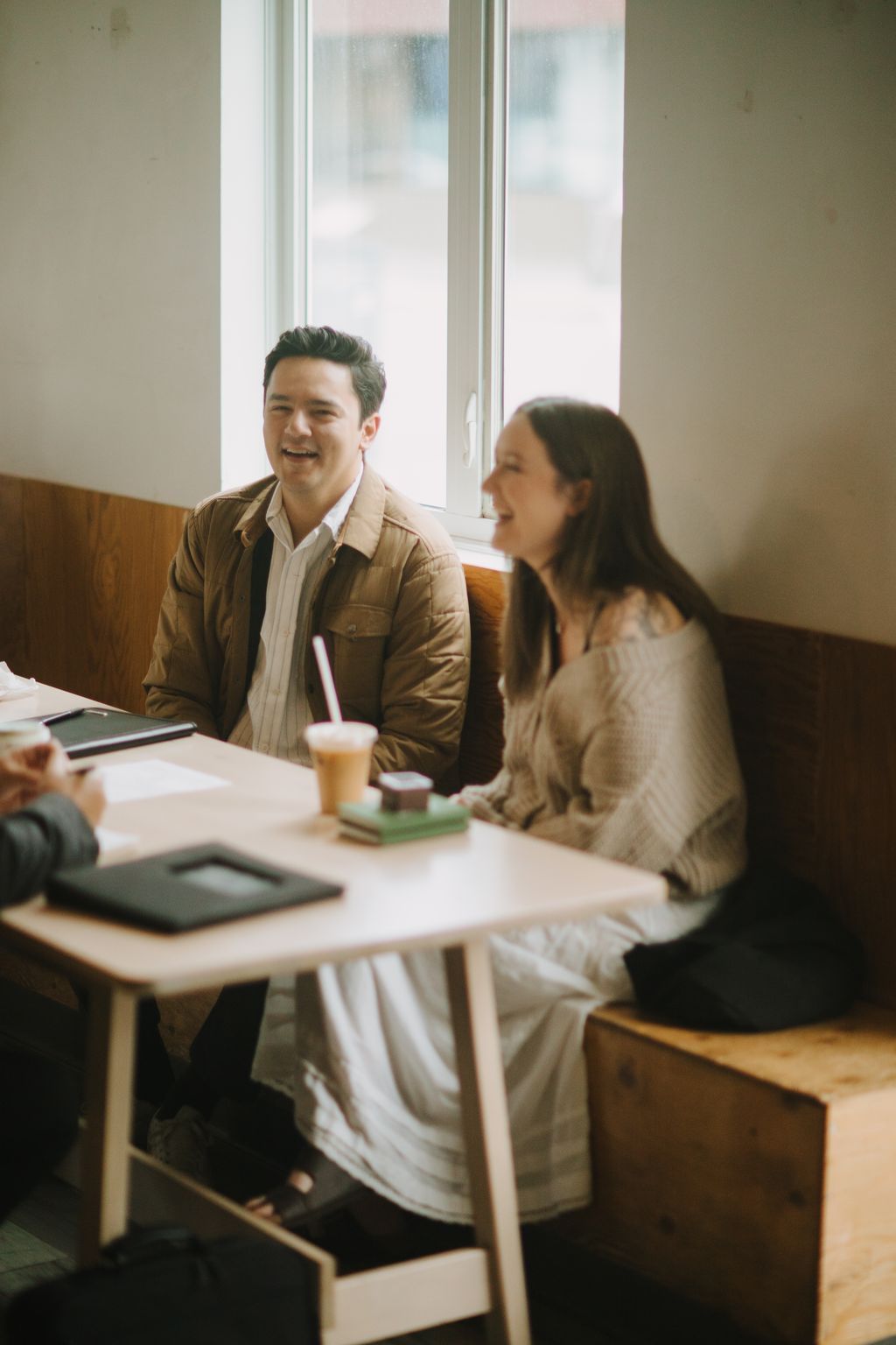 Couple laughing together by bright windows at a café, holding iced beverages in warm-toned casual clothing