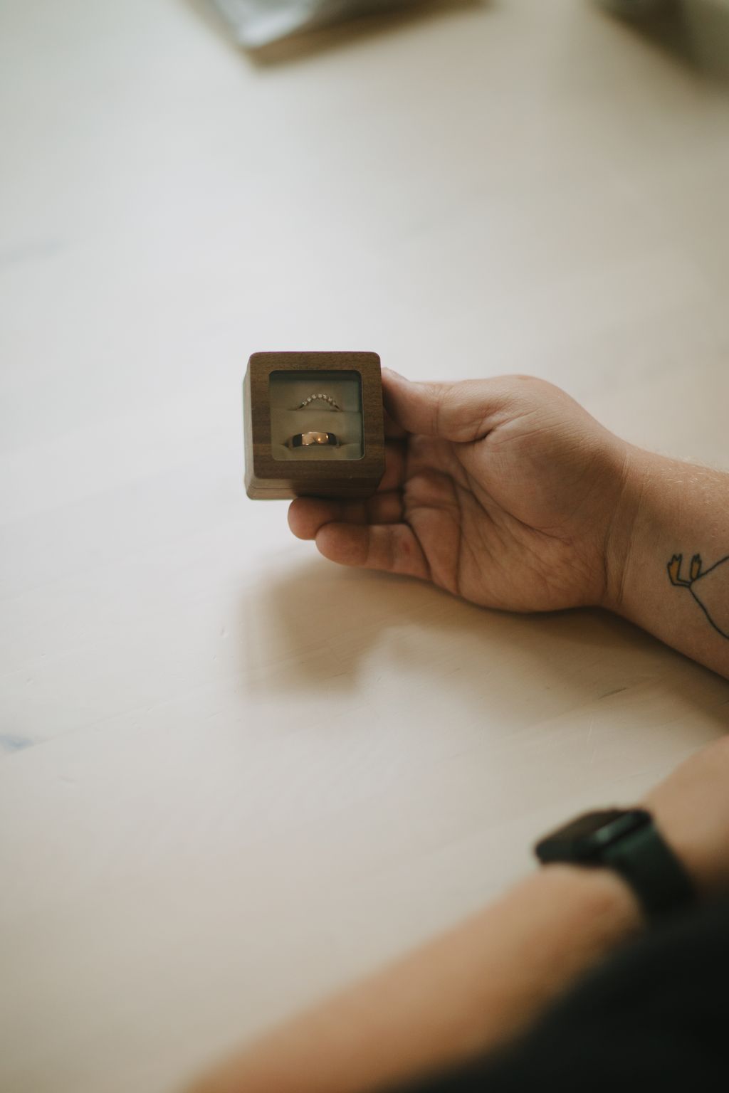 Hand holding wooden ring box displaying two gold wedding bands against neutral background