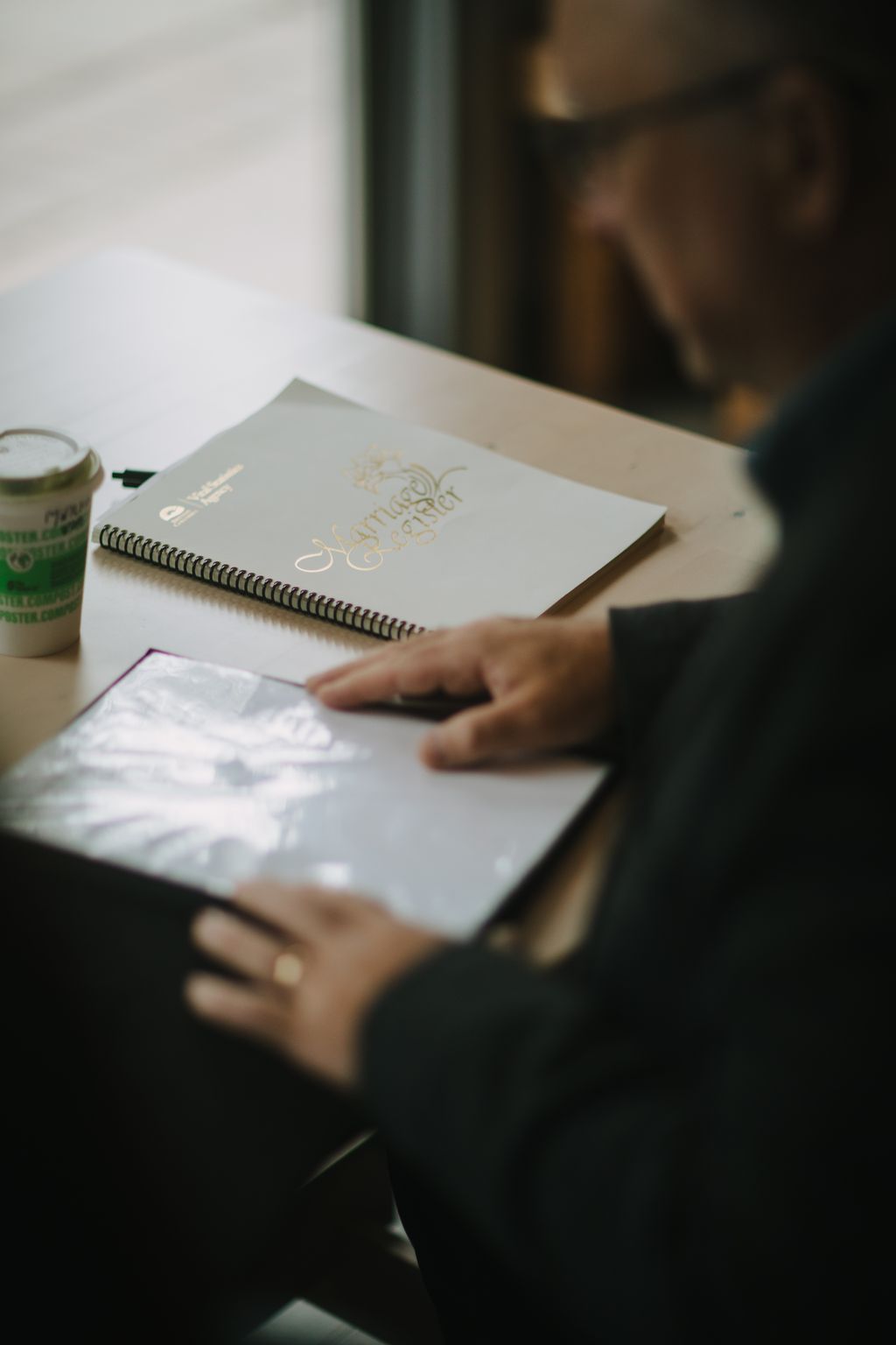 Bride and groom''s hands holding open a spiral-bound wedding guest book with gold calligraphy, soft natural light streaming across white pages