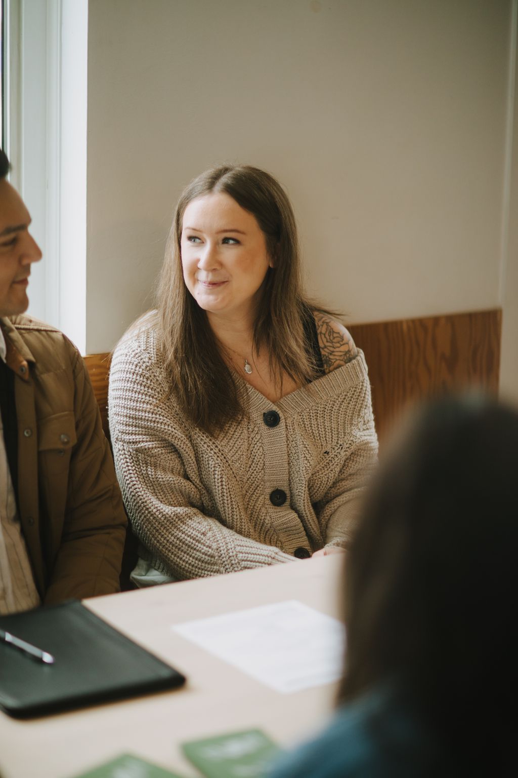 Woman in tan cardigan with tattoo smiles warmly during indoor consultation, soft natural light