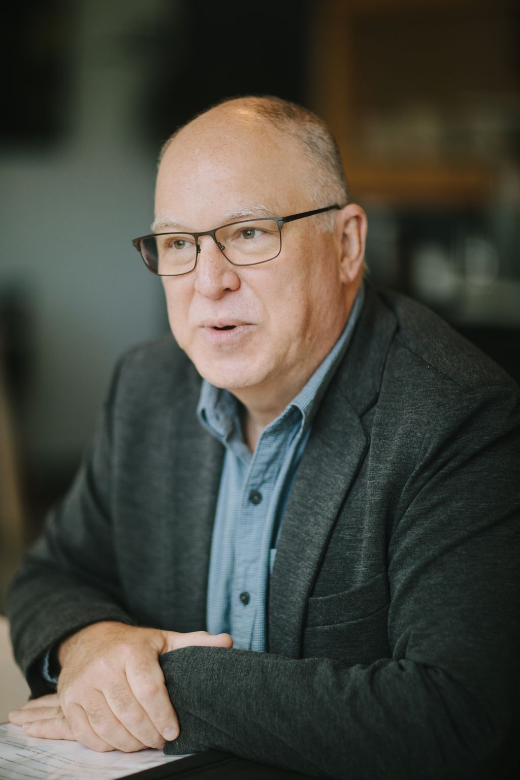 Professional headshot of a man wearing glasses and a gray blazer over a light blue checkered shirt, photographed against a blurred background