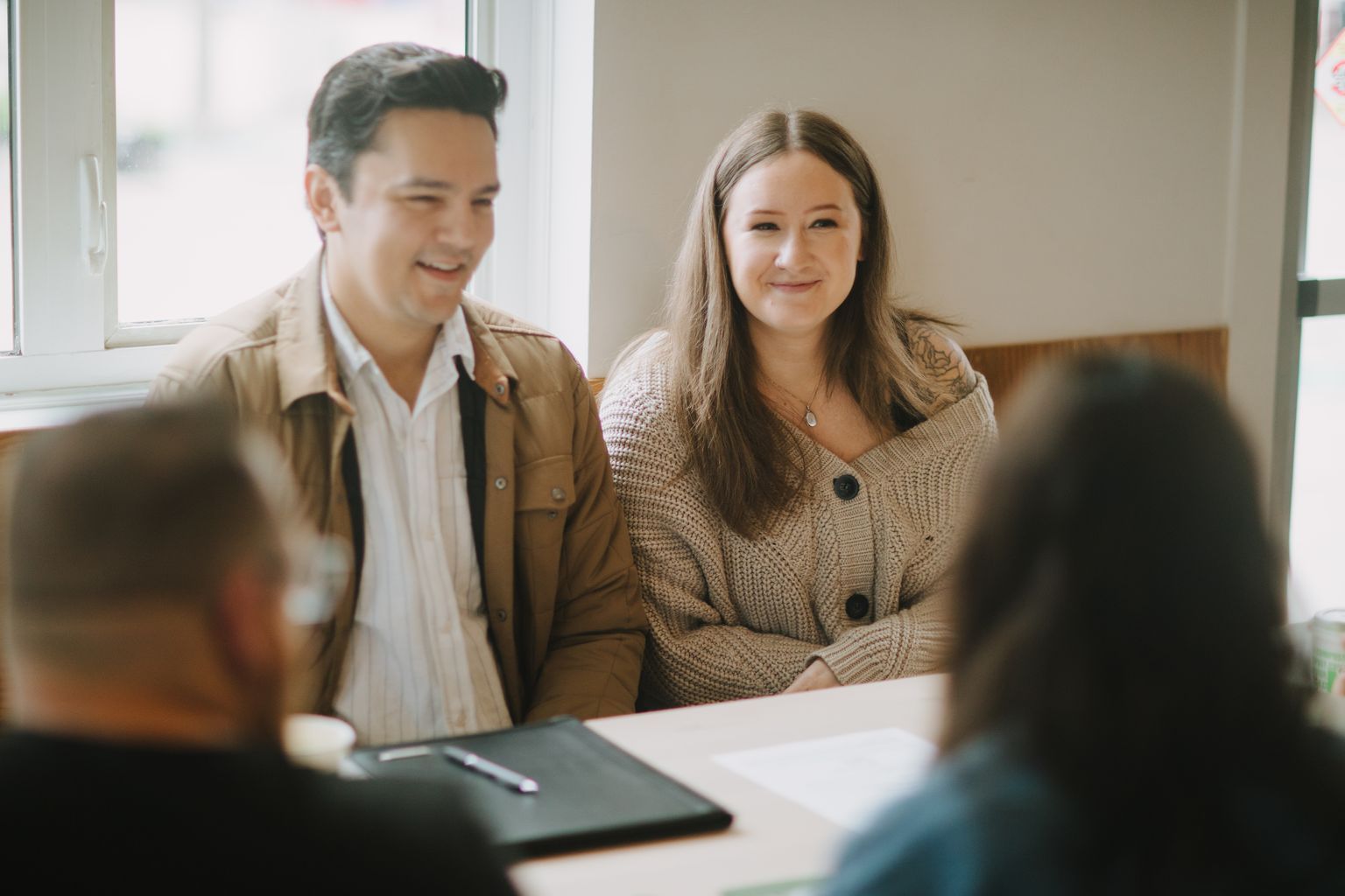 Couple smiling at desk during bright indoor engagement session, woman in beige cardigan, man in tan jacket
