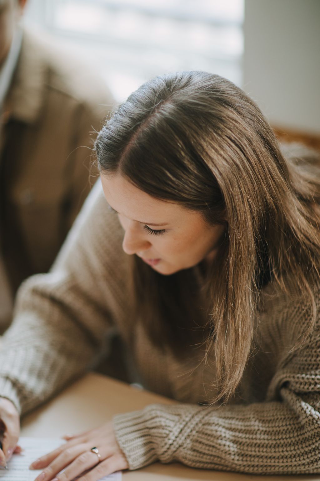Young woman with long brown hair in an updo, gazing downward in soft natural light, wearing a neutral knit sweater