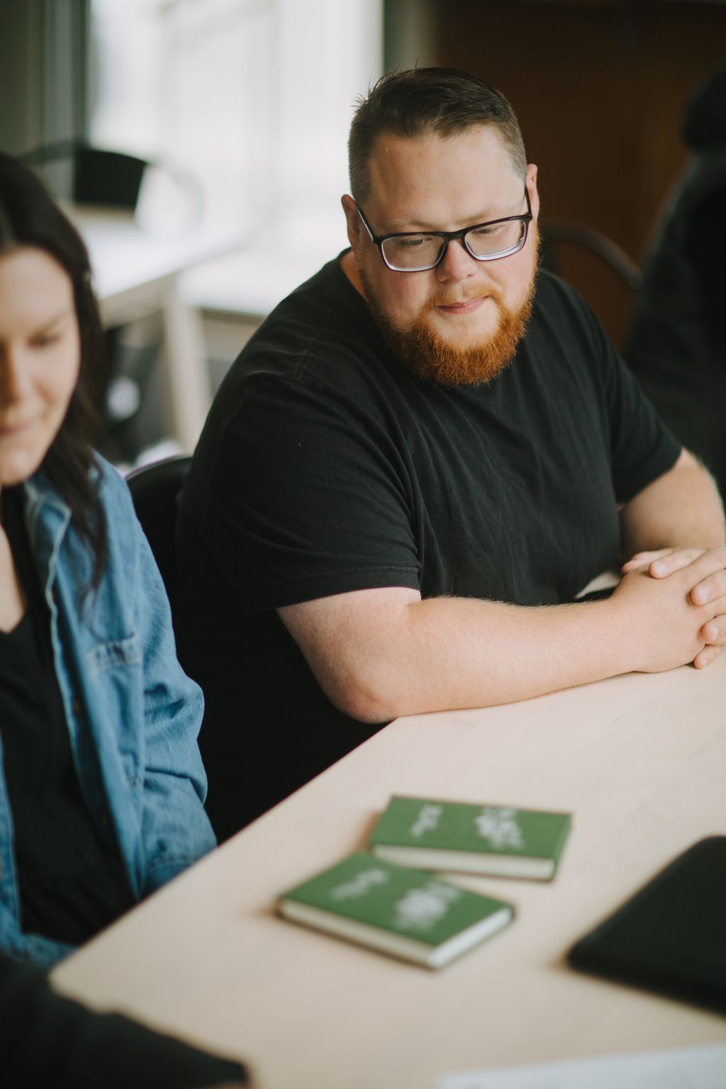 Man with beard and glasses in black shirt looking down at green book on wooden table, soft natural light