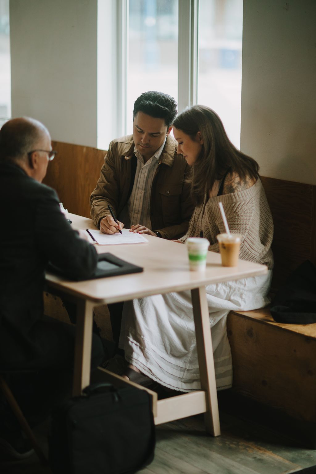 Couple reviewing wedding documents at a bright window, natural light illuminating their focused expressions
