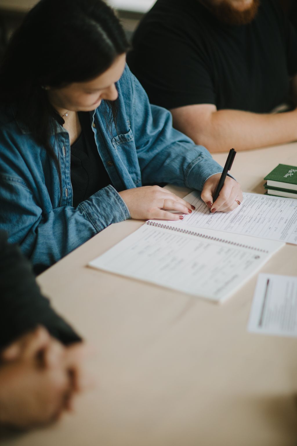 Child in denim shirt writing in notebook while seated next to adult in dark clothing at wooden table