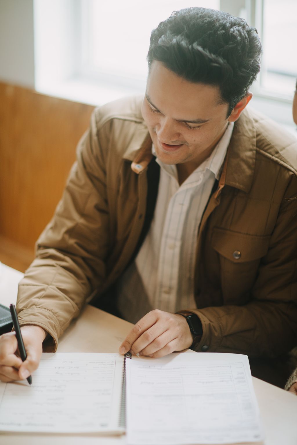 Groom in tan jacket and white shirt examines wedding details with gentle focus, natural light highlighting intimate moment