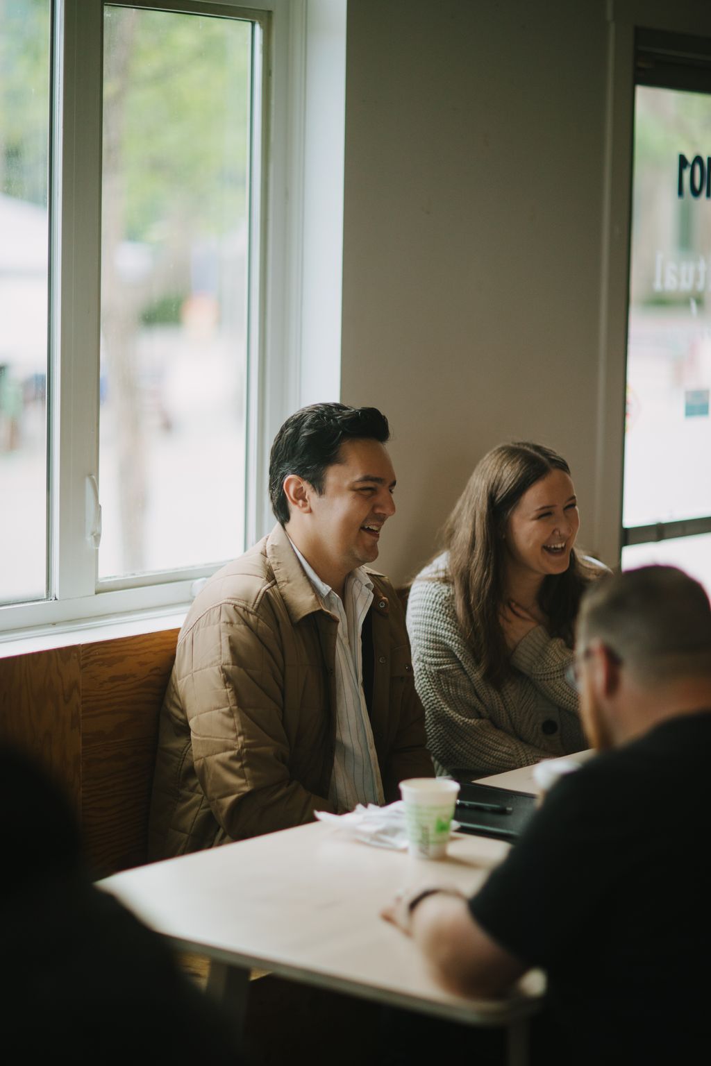 Man in tan jacket laughing during indoor gathering by bright window with natural light