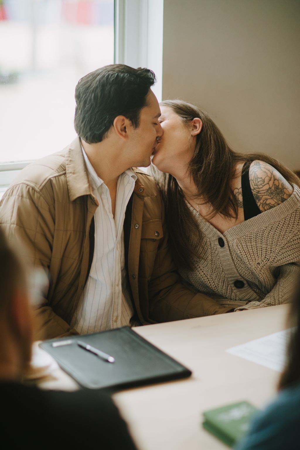 Couple kissing at desk with laptop and natural window light, intimate moment in bright indoor setting