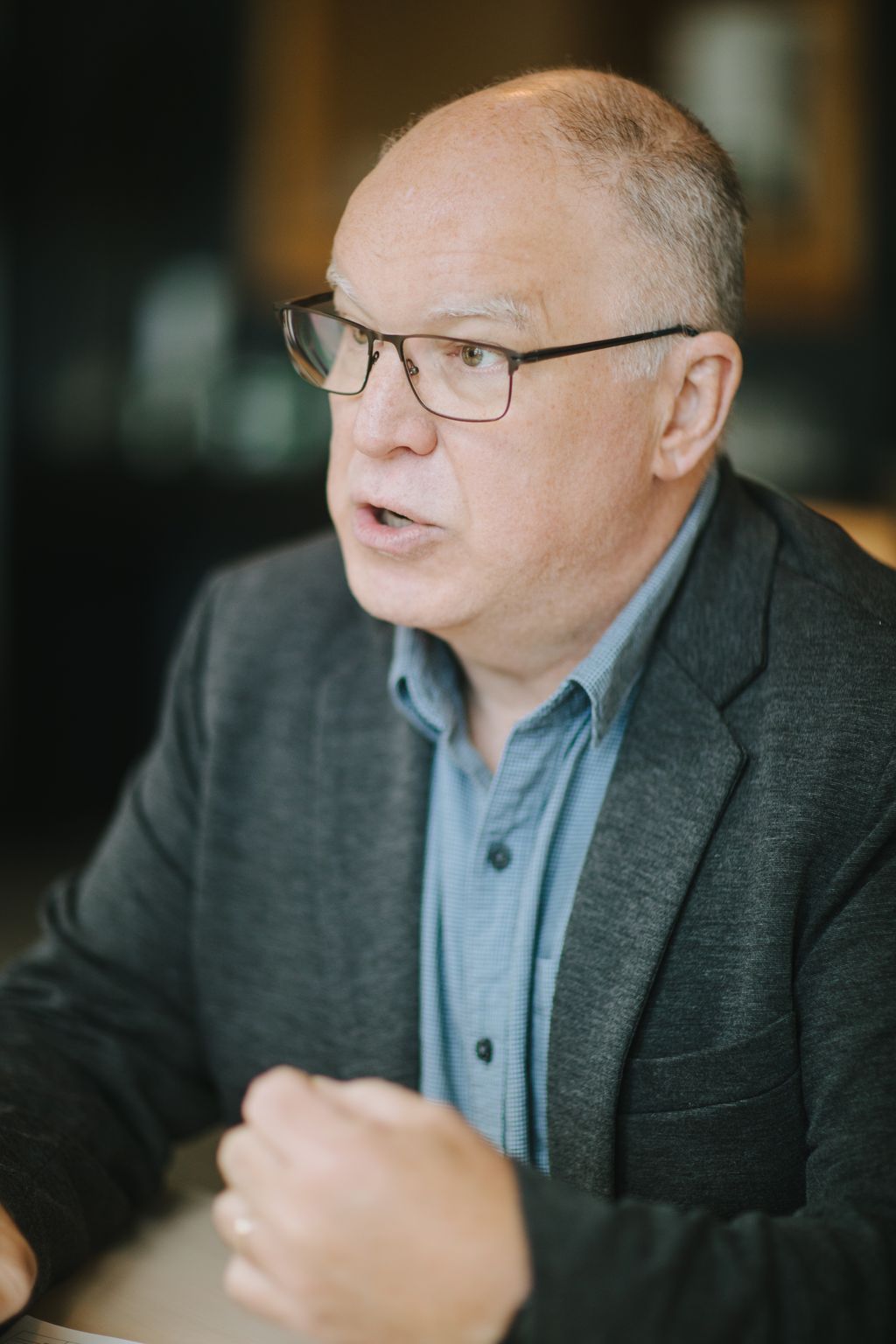 Bald man wearing glasses and light blue shirt speaks during wedding ceremony with warm, blurred background