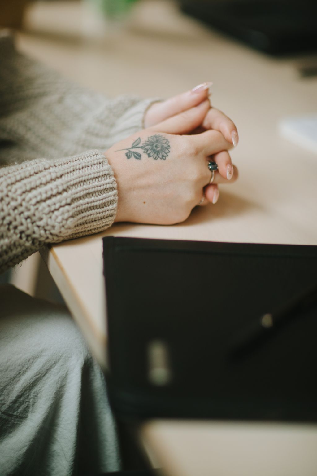 Close-up of a bride''s tattooed hand with delicate sunflower design, wearing an emerald ring and cream knit sleeve