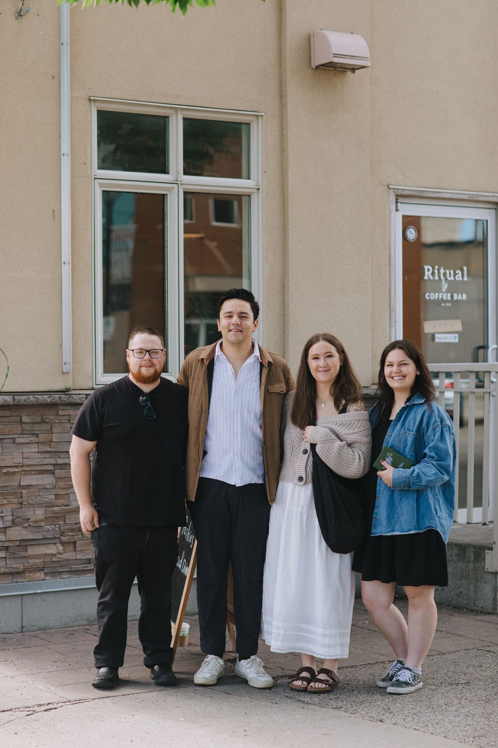 Three people standing together against a beige building wall with large windows, smiling at the camera in natural daylight