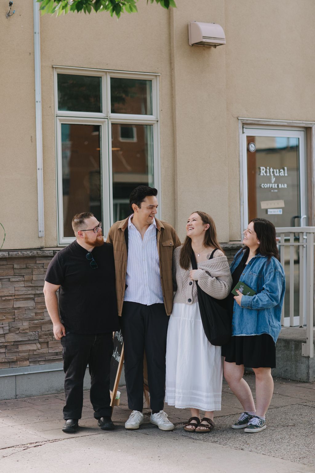 Couple smiling together outside a modern building, surrounded by friends in casual outdoor setting with warm natural light