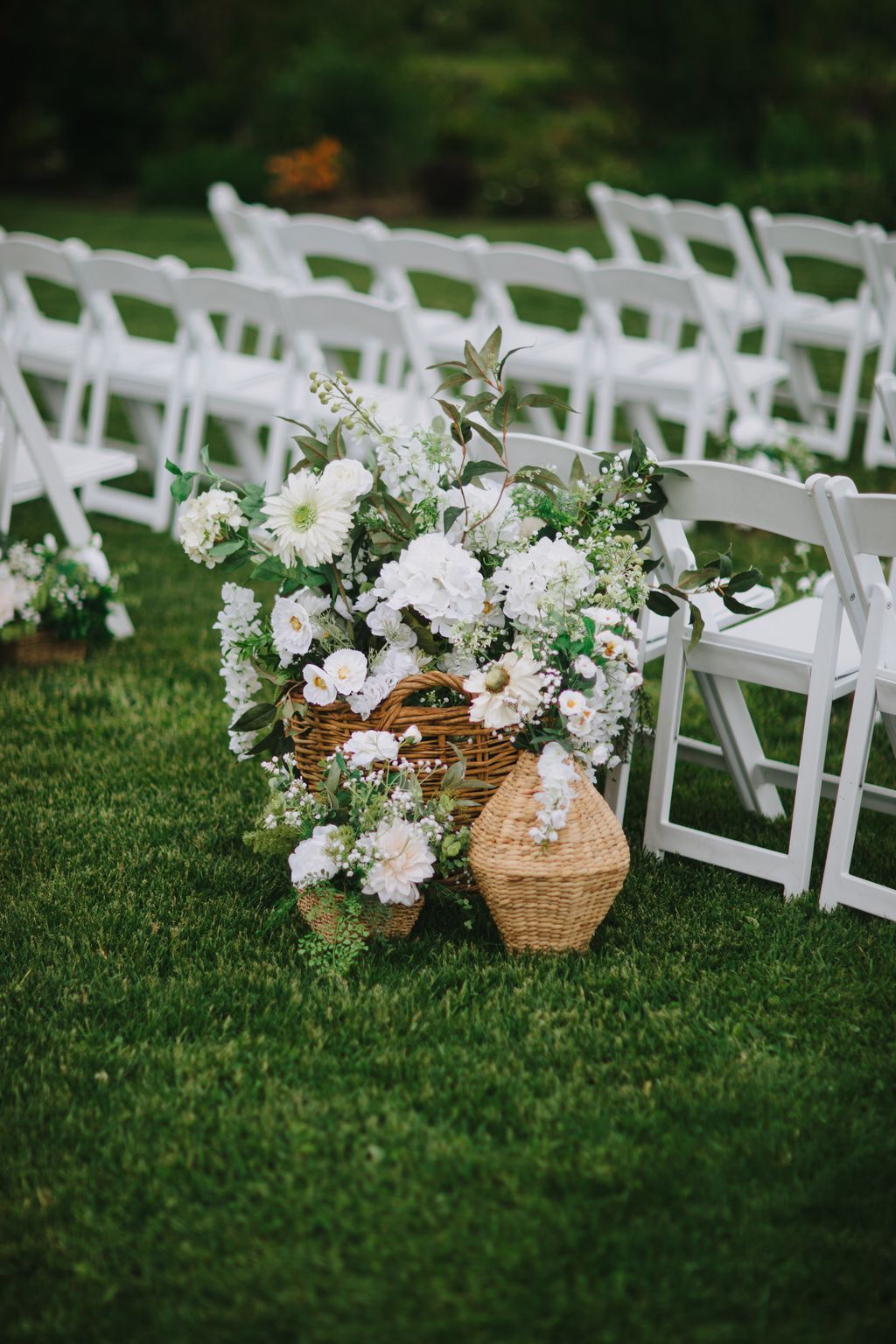 White wicker baskets overflowing with white flowers and greenery sit on grass beside a white lattice fence at an outdoor wedding venue