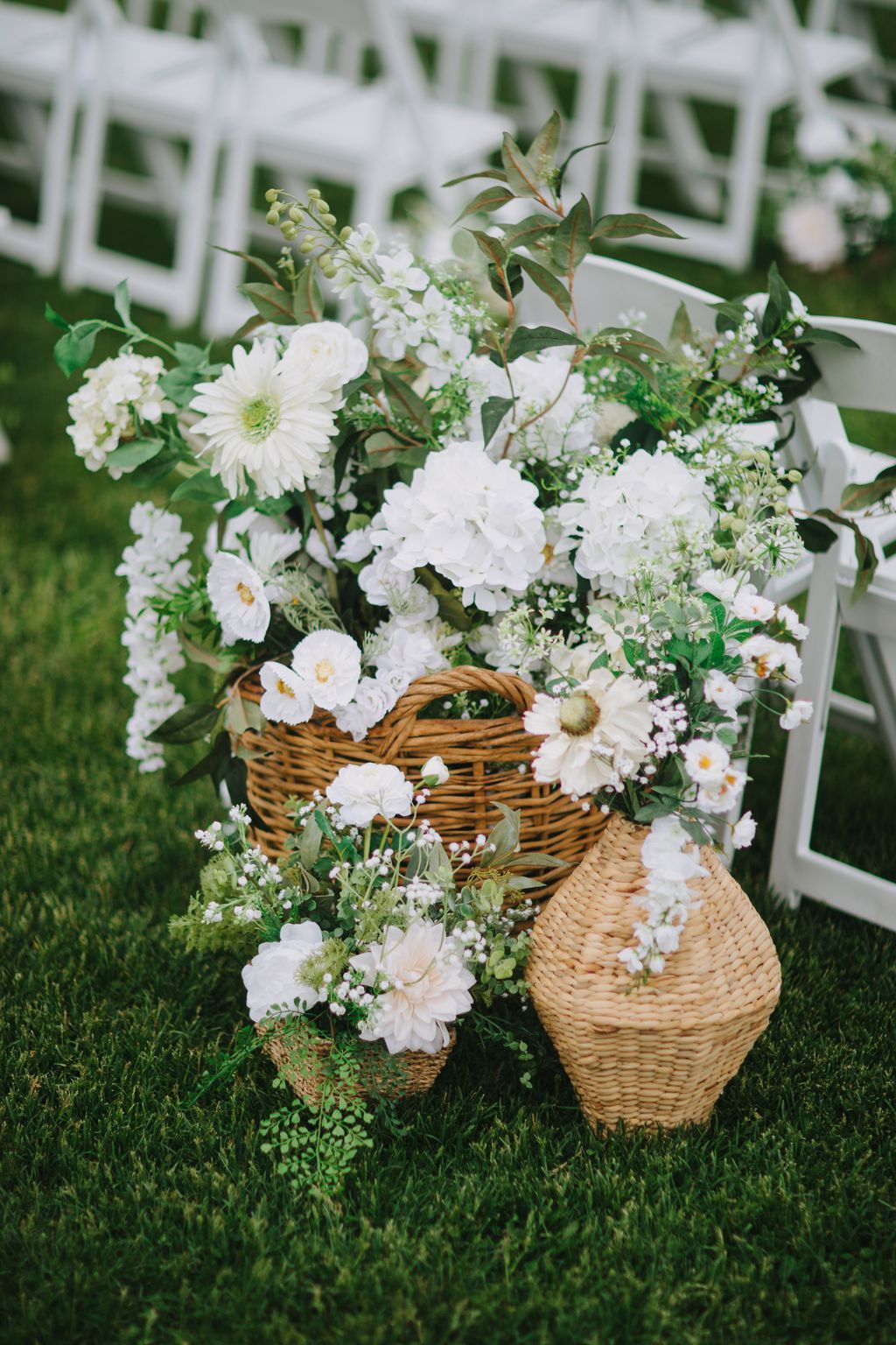 Woven baskets overflow with white flowers and greenery on grass, soft natural light creating an elegant garden wedding display