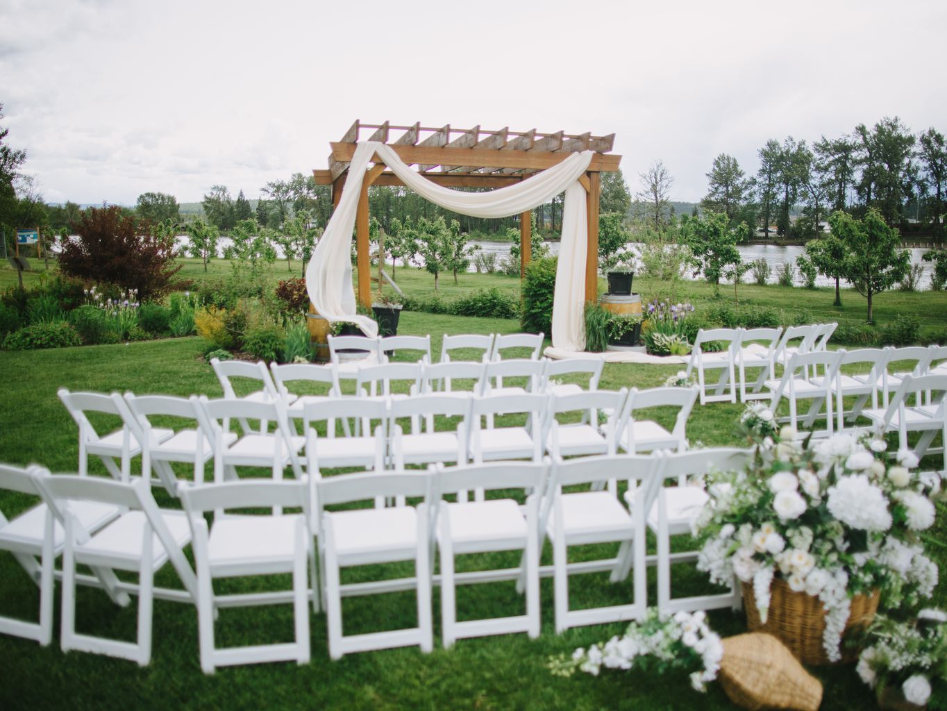 Outdoor wedding ceremony setup with wooden pergola draped in flowing white fabric and white folding chairs arranged on a manicured lawn