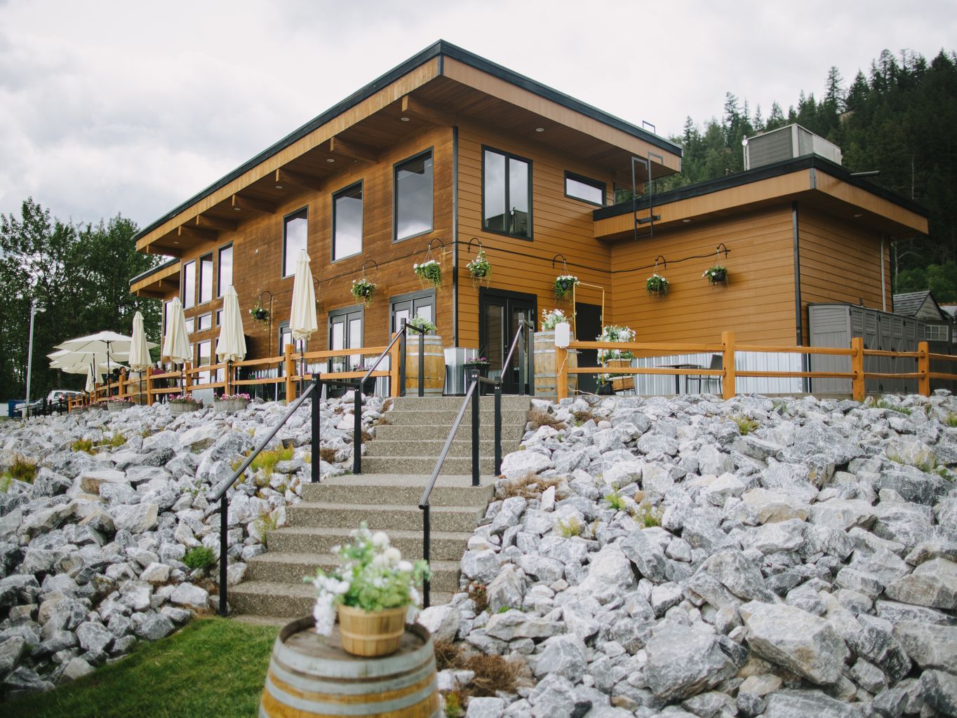 Modern wood-clad venue with stone steps, black railings, and hanging floral arrangements under overcast sky