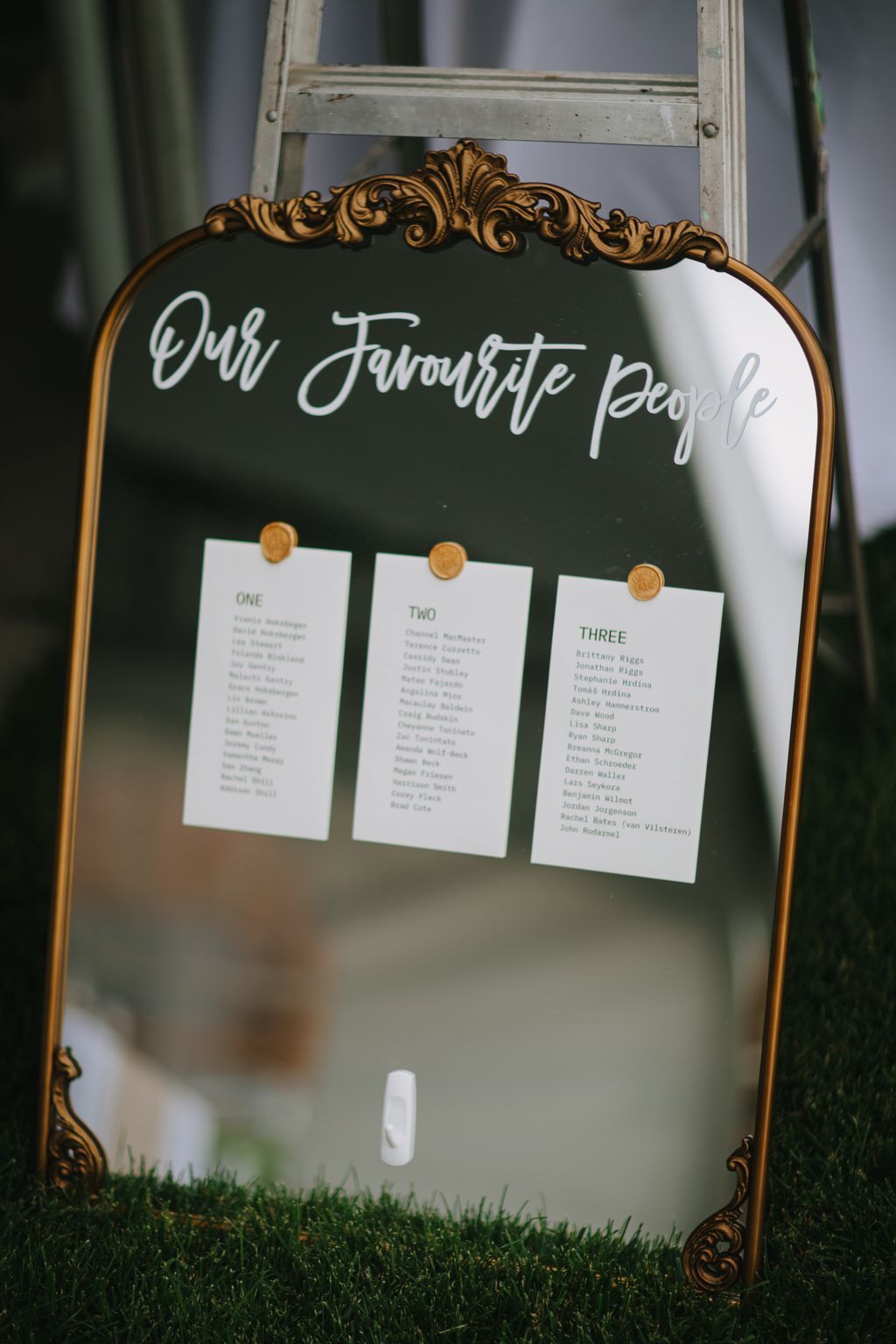 Green chalkboard seating chart with ornate gold trim displaying guest names in three numbered sections, hung with gold clothespins