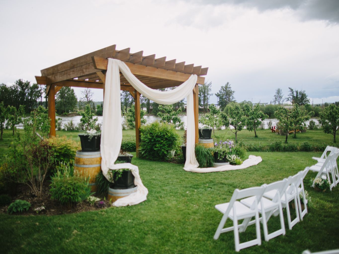 Wooden pergola with flowing white fabric draping and potted plants on manicured lawn, waterside backdrop