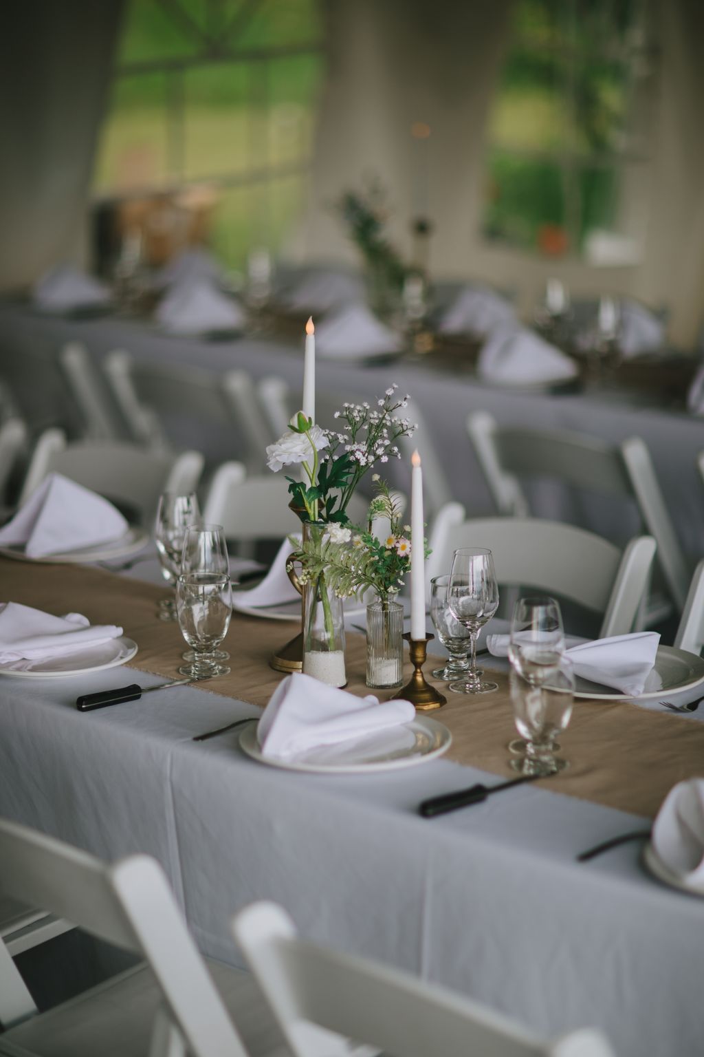 Elegant outdoor wedding tablescape with white linens, lit candles in brass holders, fresh white flowers in glass vases, and place settings on a wooden table