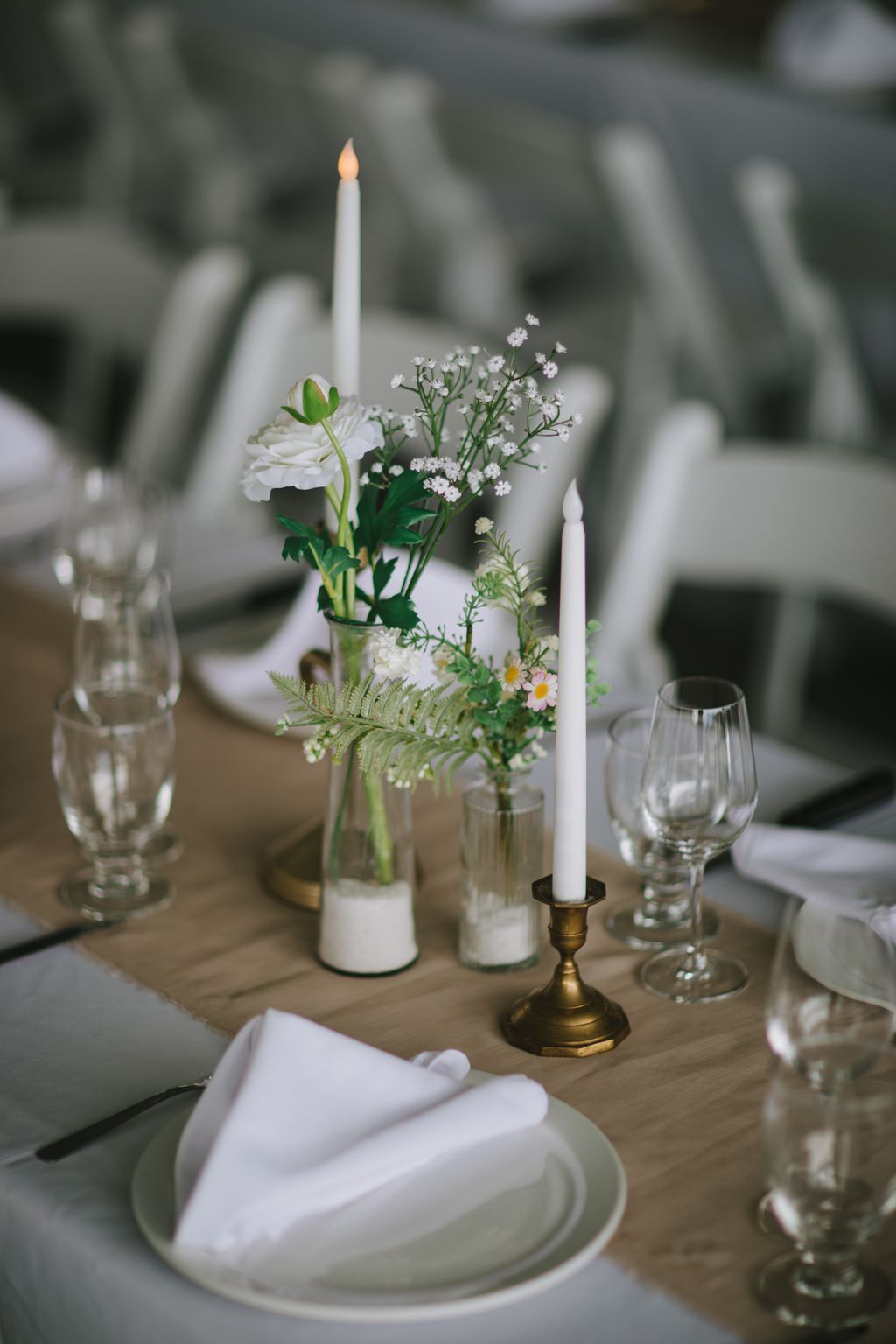 Elegant wedding table setting with white flowers, baby''s breath, and fern in glass vases flanked by lit white taper candles in brass holder