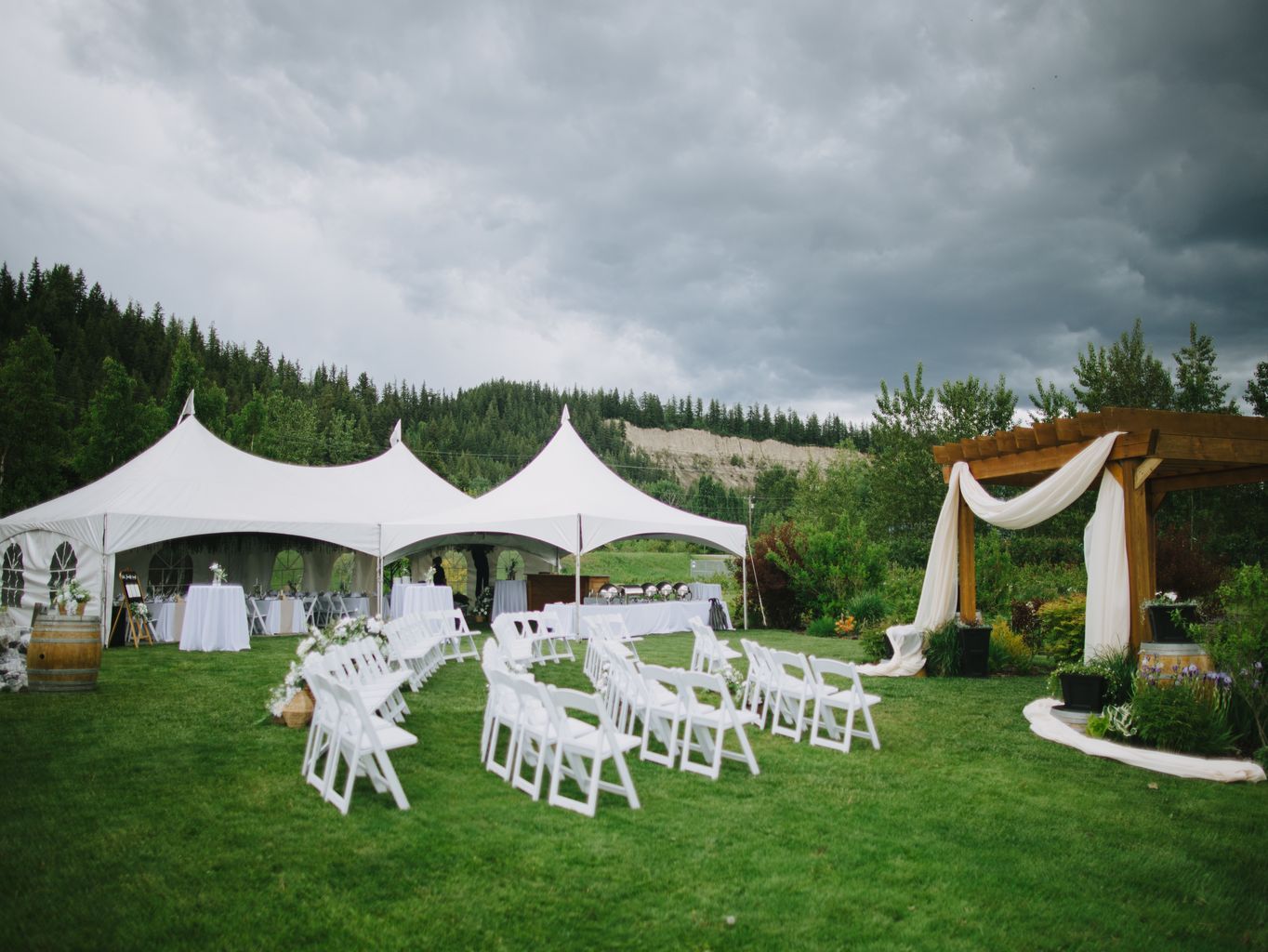White folding chairs arranged in semicircles on manicured lawn facing elegant tent with forested mountains under overcast sky