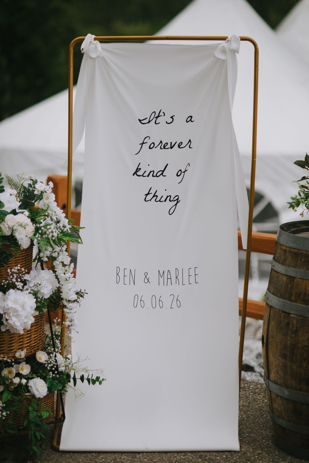 White fabric banner hung from wooden rod with handwritten text "it''s a forever kind of thing" and couple''s names and wedding date, greenery visible at edges