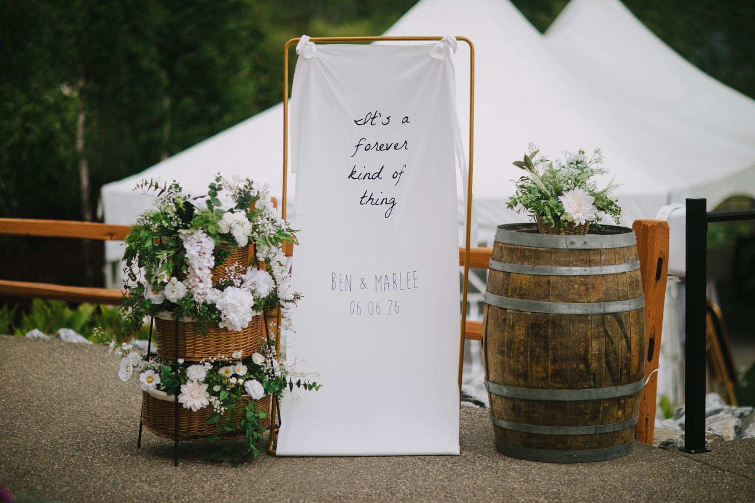 White fabric banner with handwritten text "it''s a forever kind of thing" and couple''s names displayed at outdoor wedding ceremony entrance
