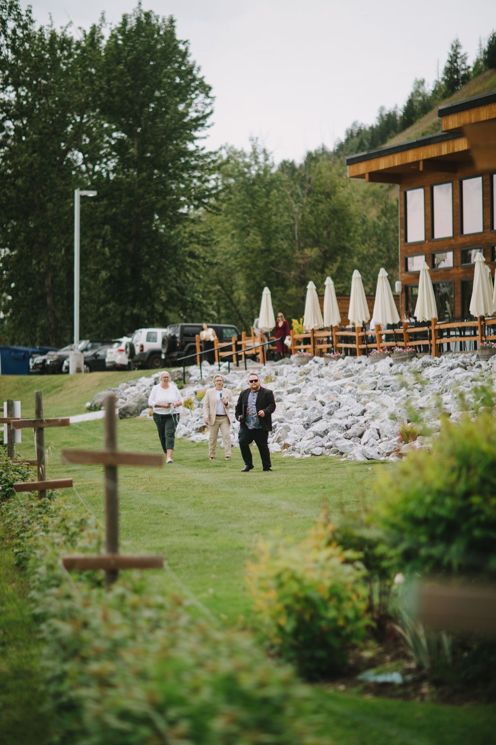 Guests walk across manicured lawn toward stone terrace with white umbrellas, forested mountains and wooden structure behind