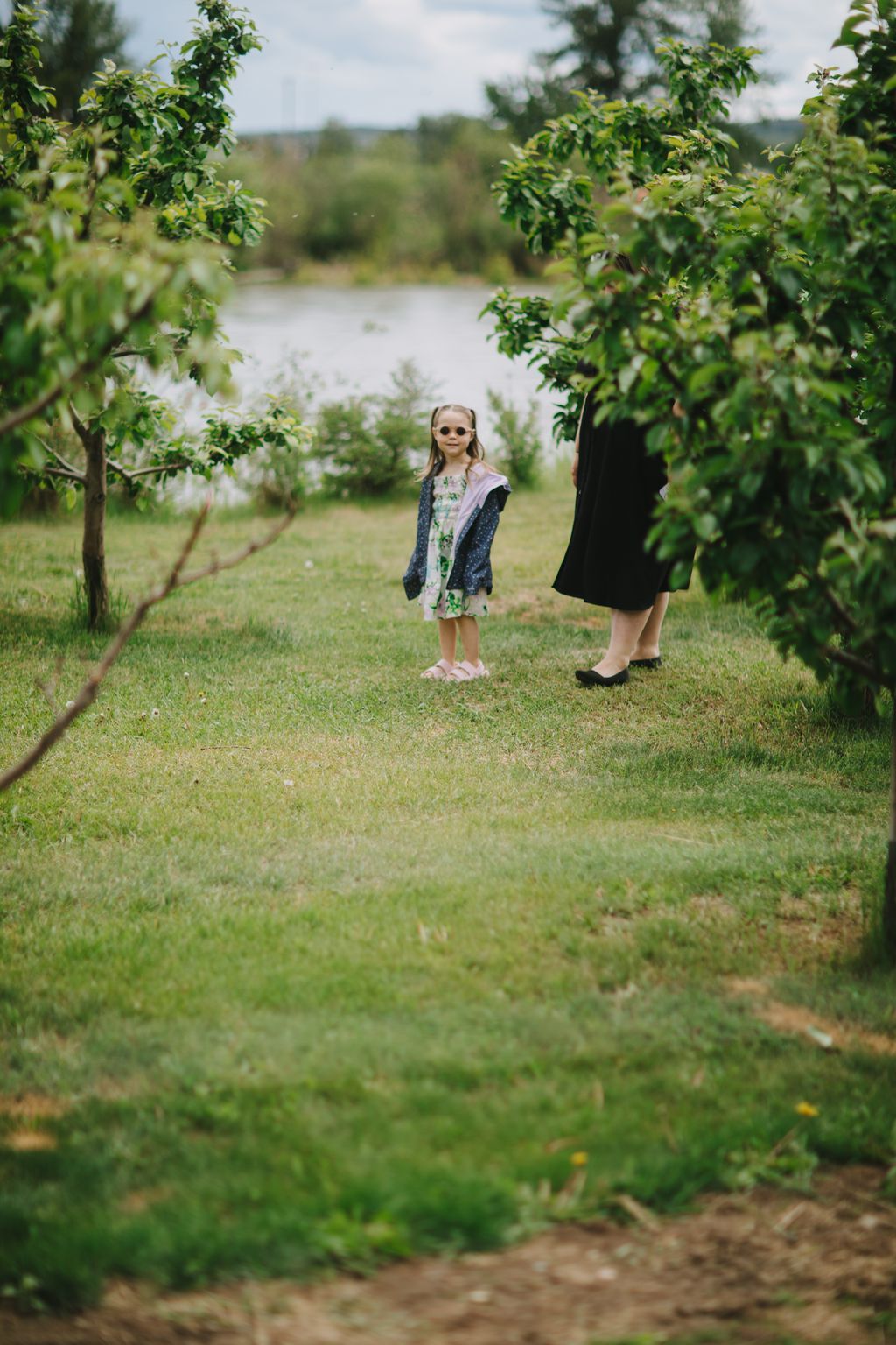 Young girl in sunglasses and floral dress stands on grass by a river, framed by green foliage and trees