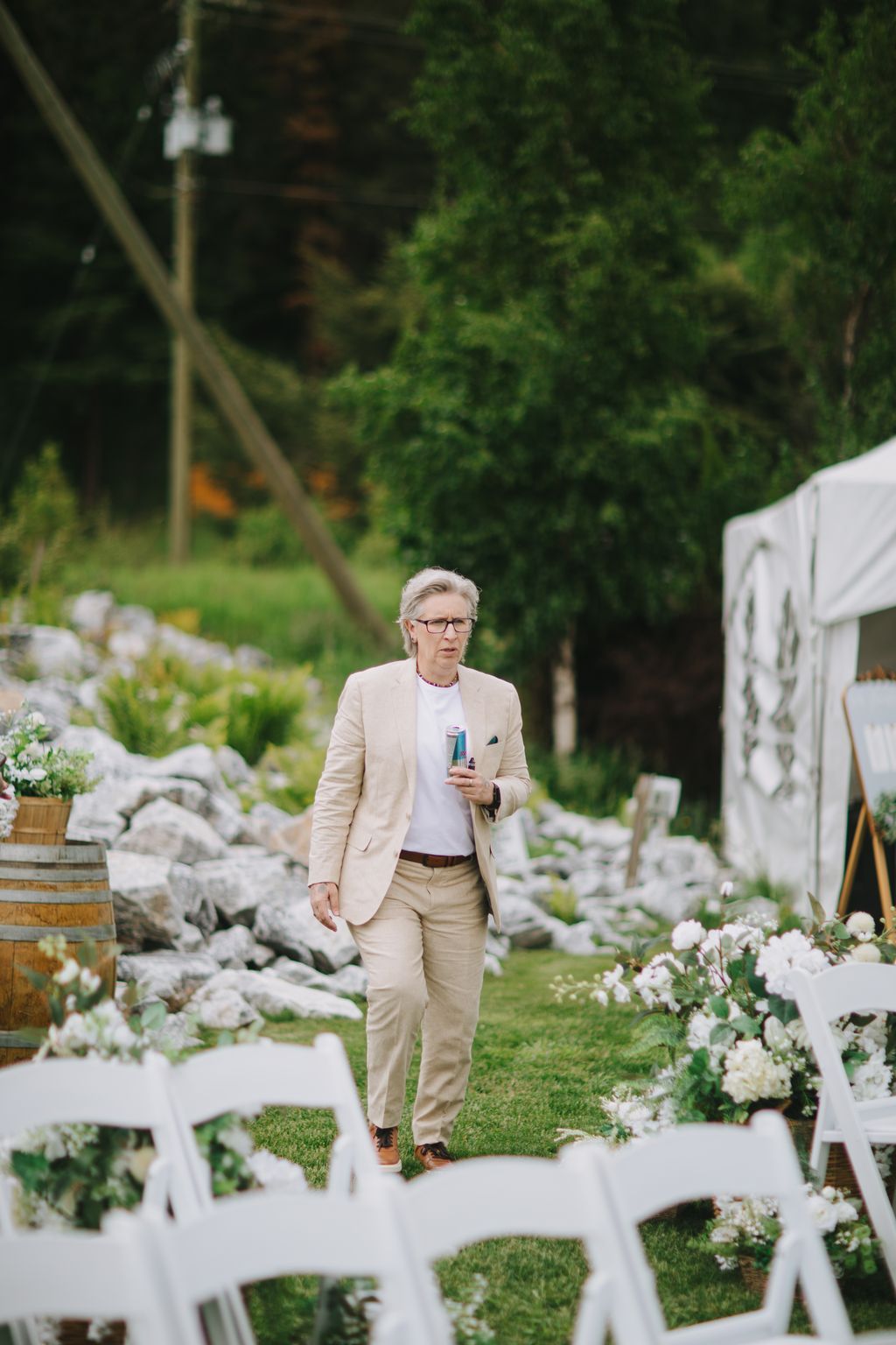 Groom in cream blazer and pants walks through lush garden pathway holding a drink, soft natural light filtering through green foliage behind him