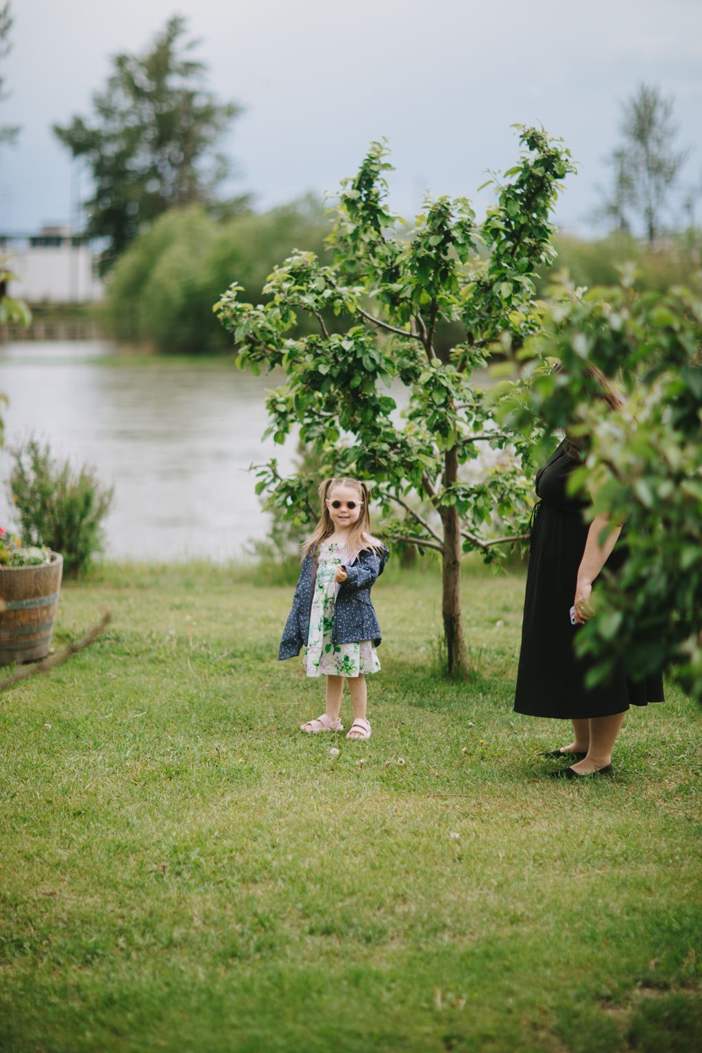 Young girl in floral dress and cardigan standing on grass beside young tree near water, wearing sunglasses