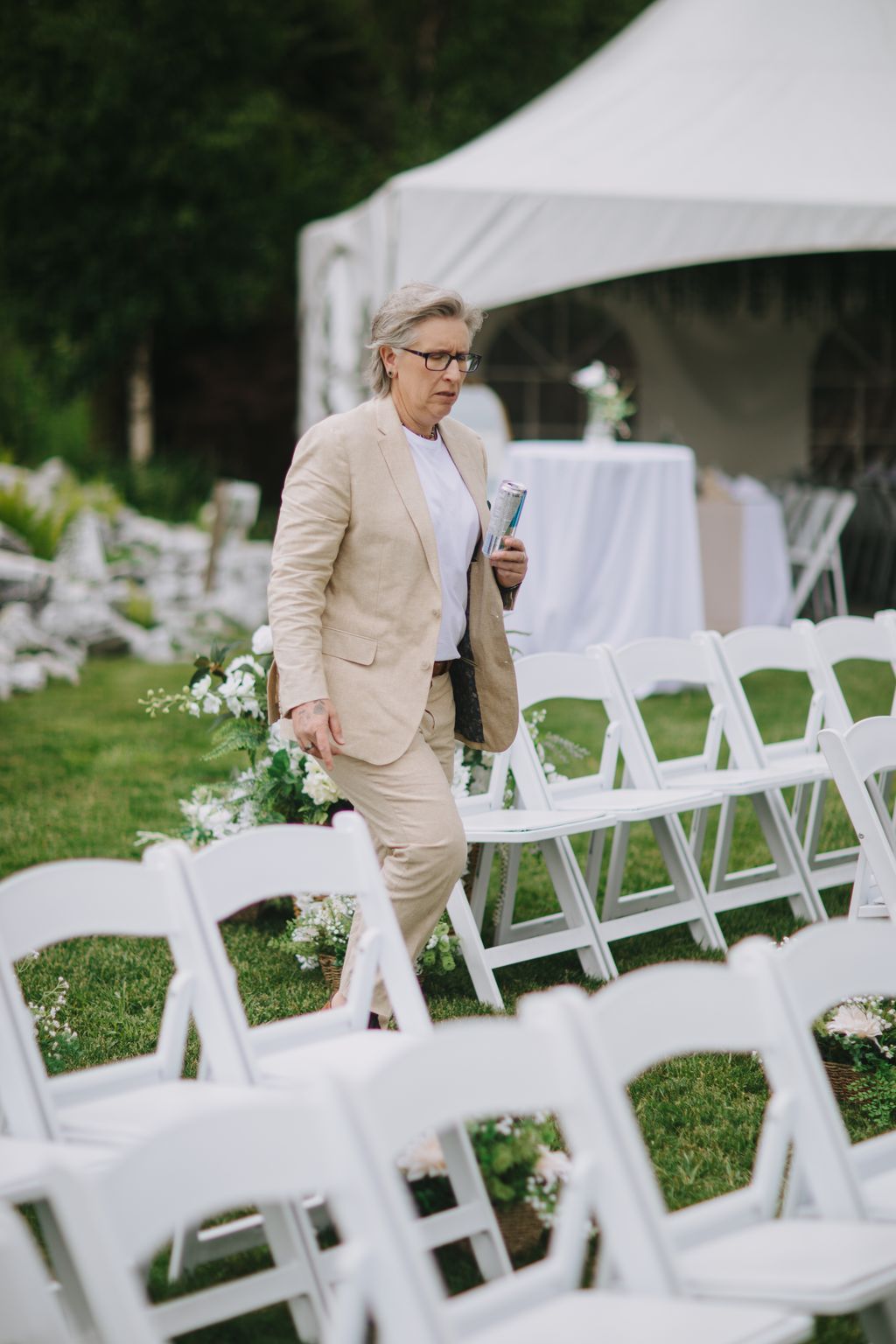Elegant older woman in beige suit holding program at outdoor garden wedding ceremony under white tent
