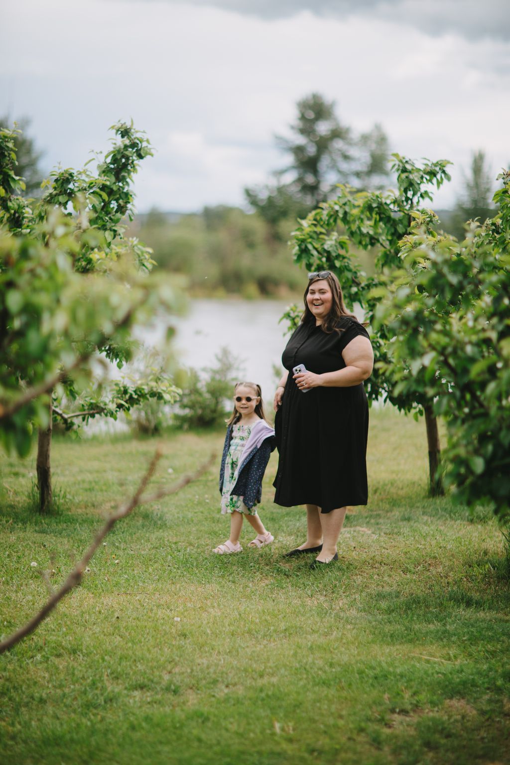 Woman and young girl holding hands on grassy riverside setting with overcast sky and green foliage