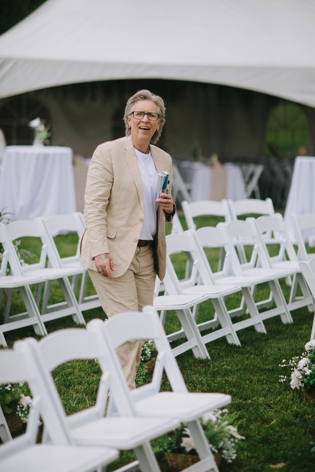 Groom in beige suit holding a drink stands among white chairs on grass with a dark tent structure behind him