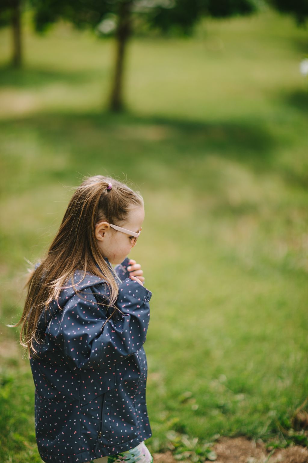 Young girl in pink glasses and navy polka dot jacket standing in soft-focus garden setting