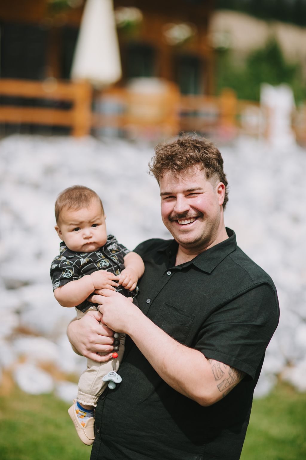 Man in black polo holding smiling toddler outdoors, blurred garden background with white umbrella
