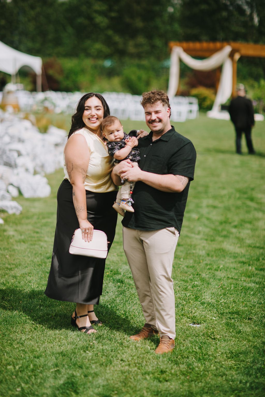Young family smiling outdoors on manicured lawn with white fence and green trees in background