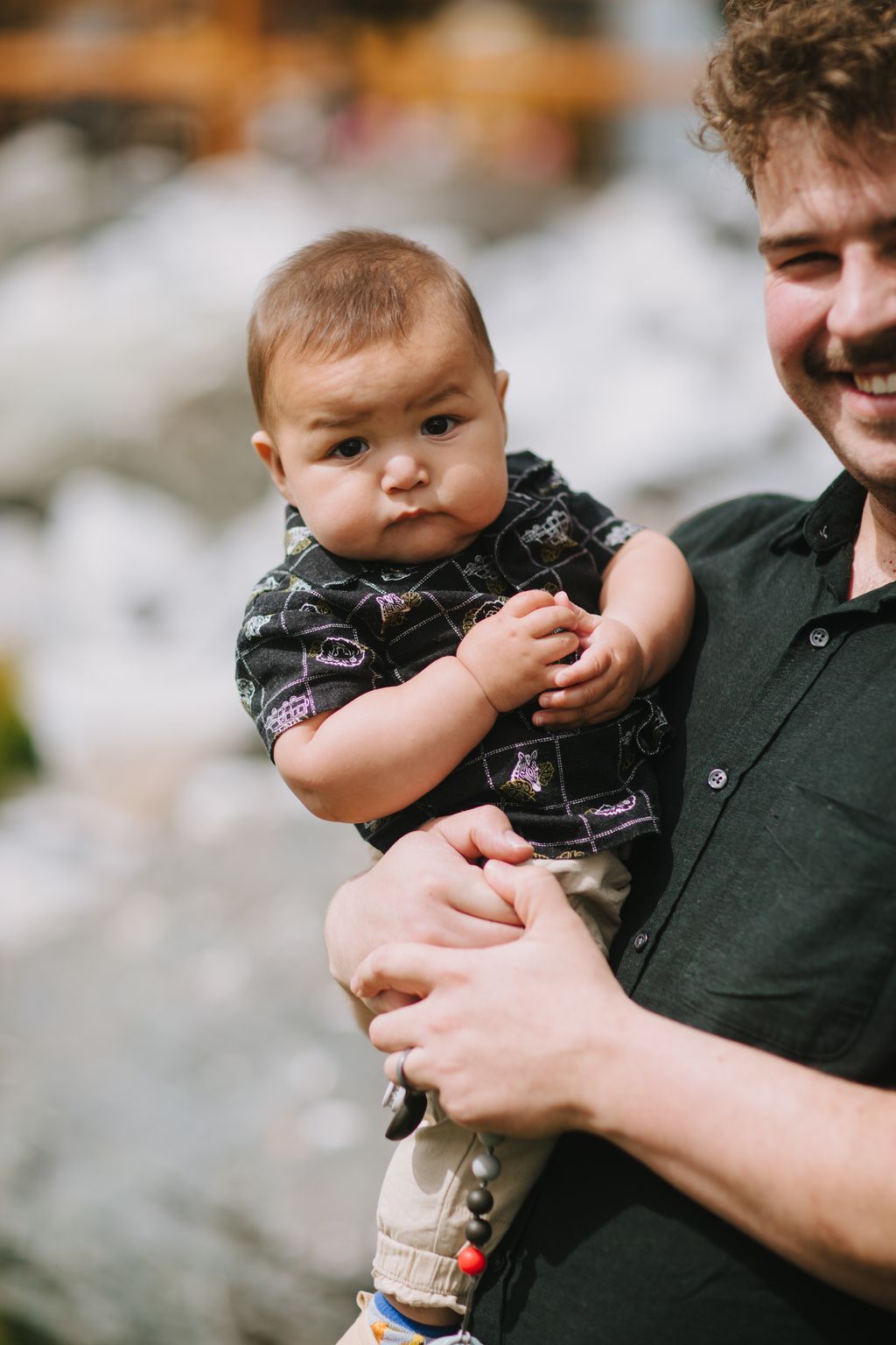 Infant in black embroidered vest held by adult, soft outdoor light, blurred background, tender family moment