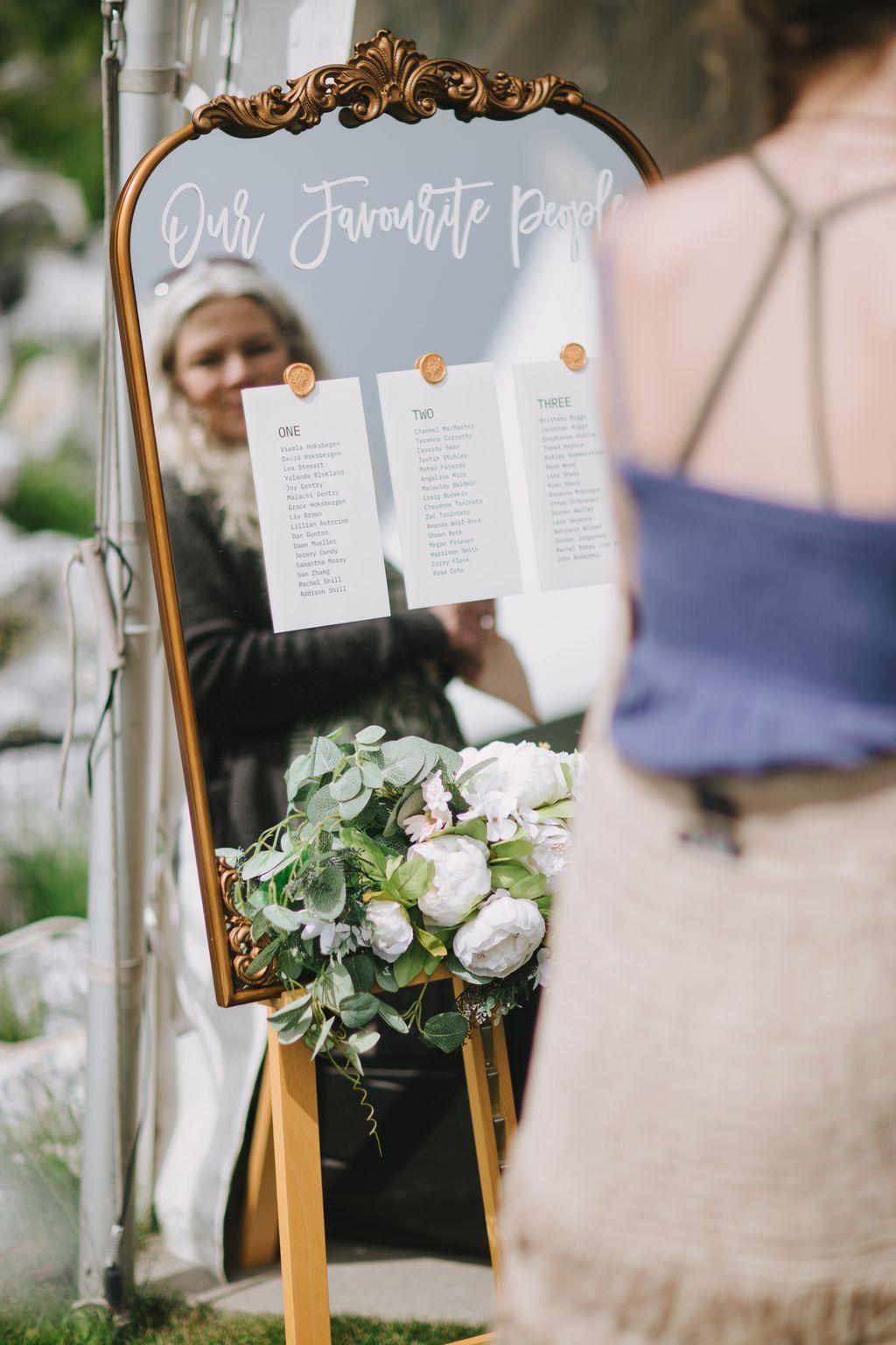 Elegant seating chart with three white cards numbered ONE, TWO, THREE held by gold wires on ornate gold frame, white peonies and eucalyptus bouquet below