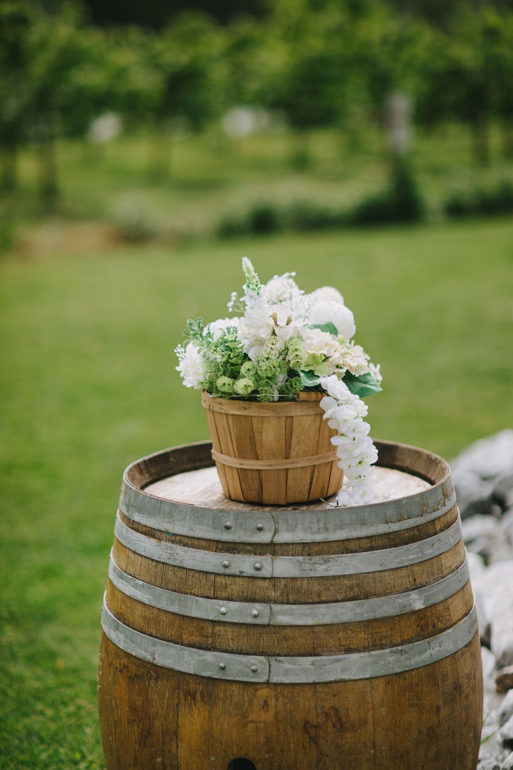 White and green floral arrangement in wooden basket atop rustic barrel, soft-focused garden backdrop