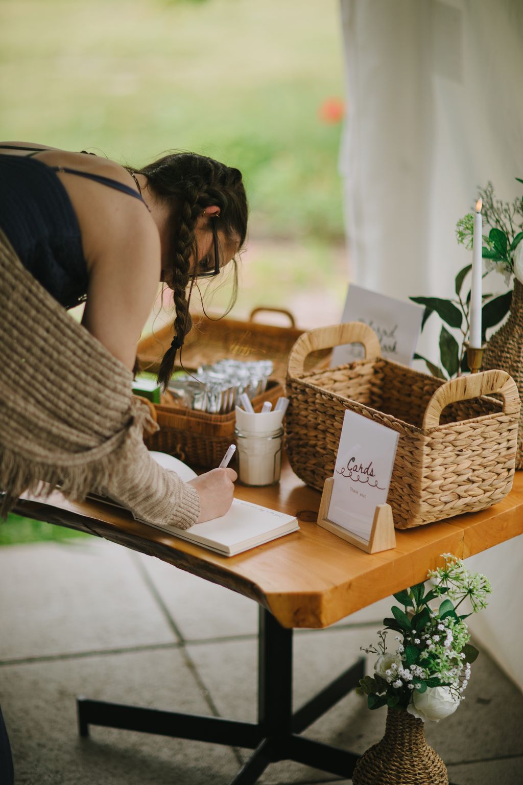 Woman with braided dark hair writing at wooden table with wicker baskets and cards, bright natural window light behind her