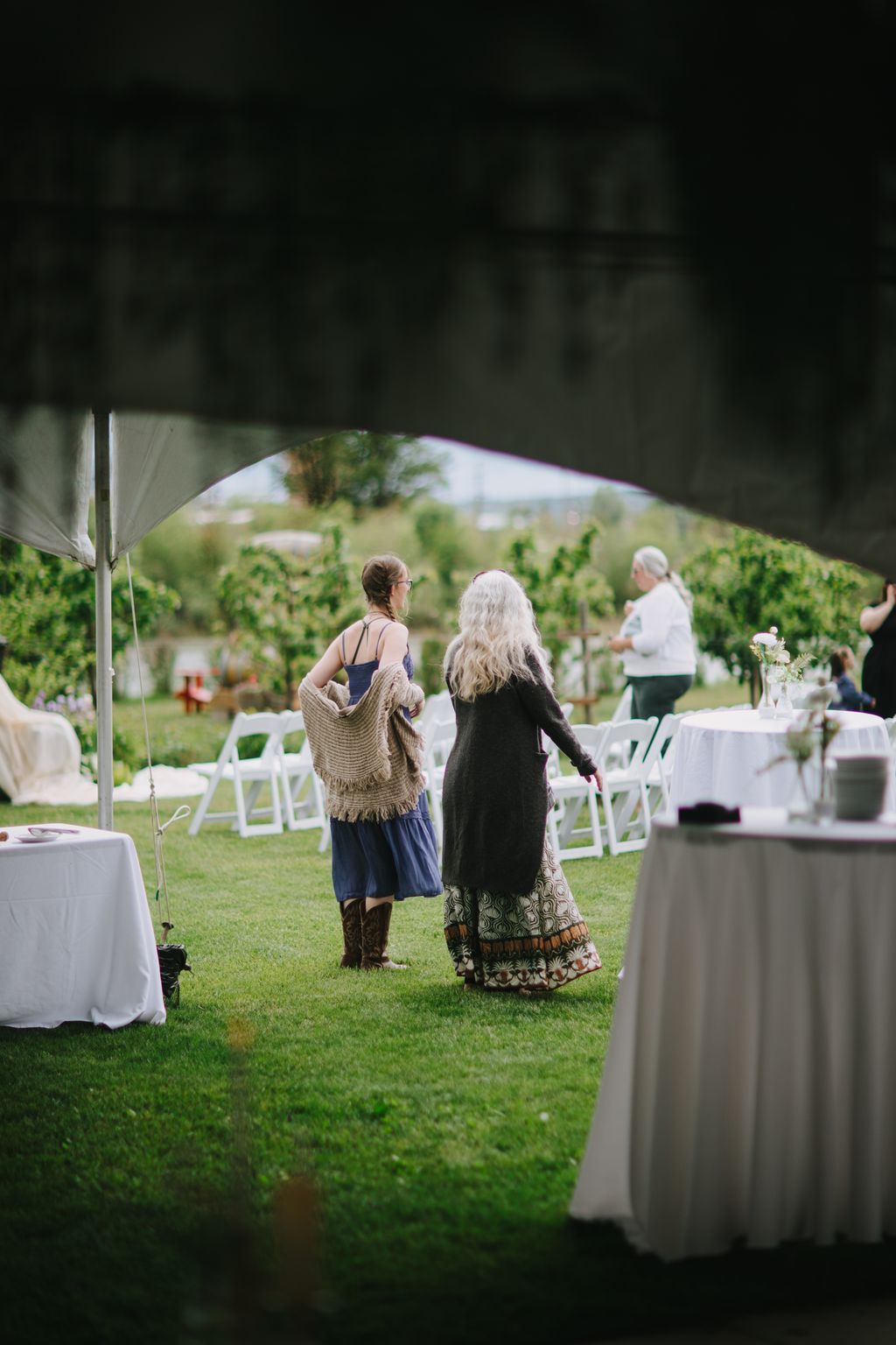Two women in elegant attire walk across a manicured lawn during an outdoor wedding ceremony, with white chairs and vineyard views visible behind them