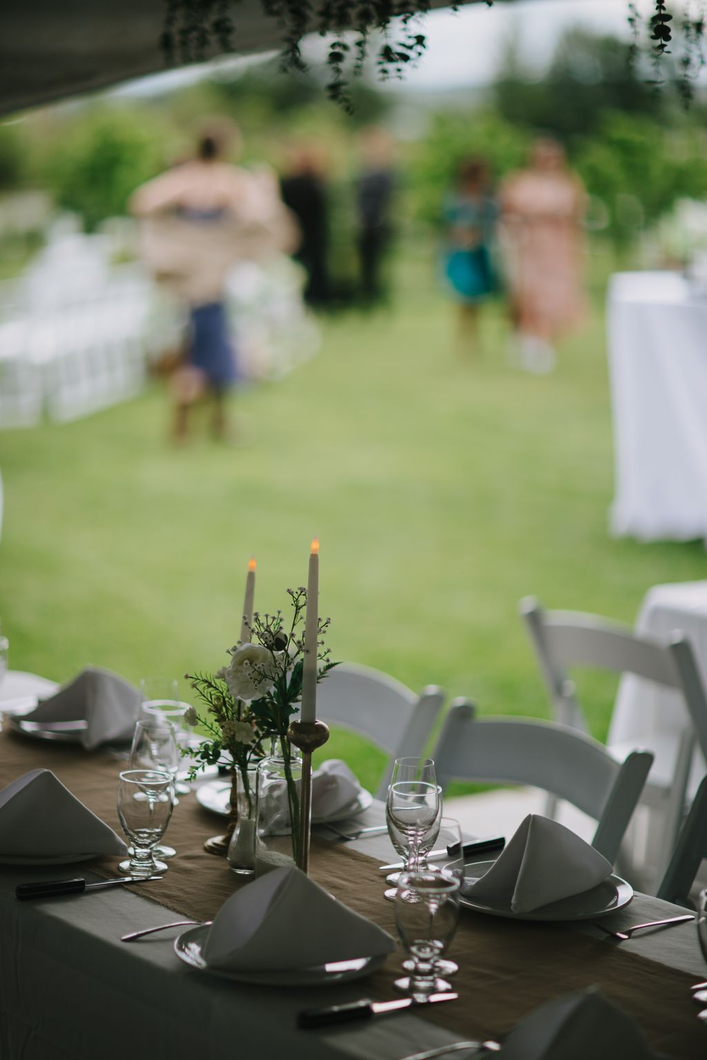 Elegant outdoor reception table with lit candles, fresh floral arrangement, and glassware, guests gathered on lawn in background