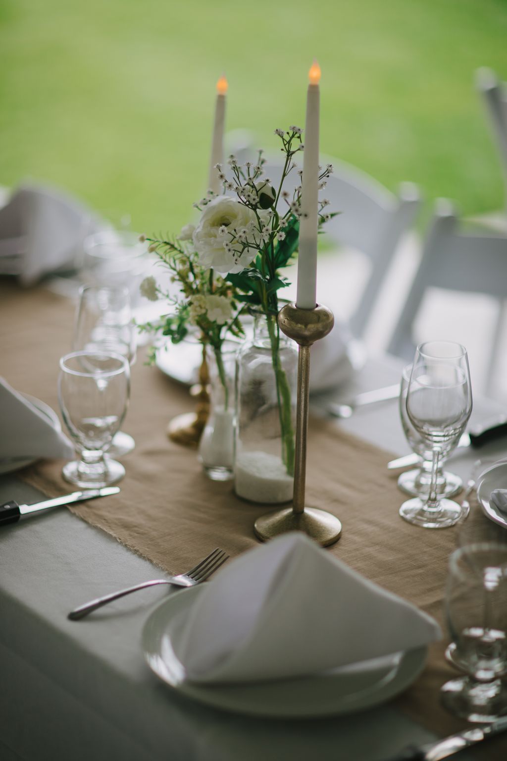 Elegant table setting with white ranunculus and baby''s breath in brass candleholder, burning candles, and folded napkin under soft natural light