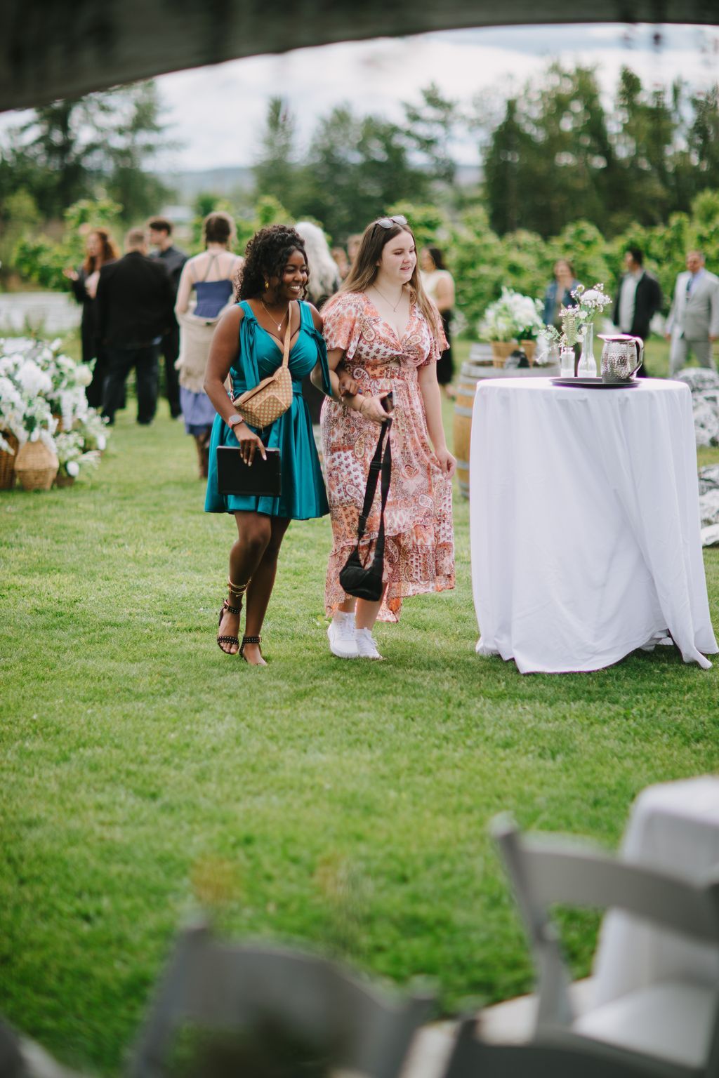 Two guests walk across a sunny garden lawn during an outdoor wedding reception, one wearing a teal dress and the other a patterned coral wrap dress
