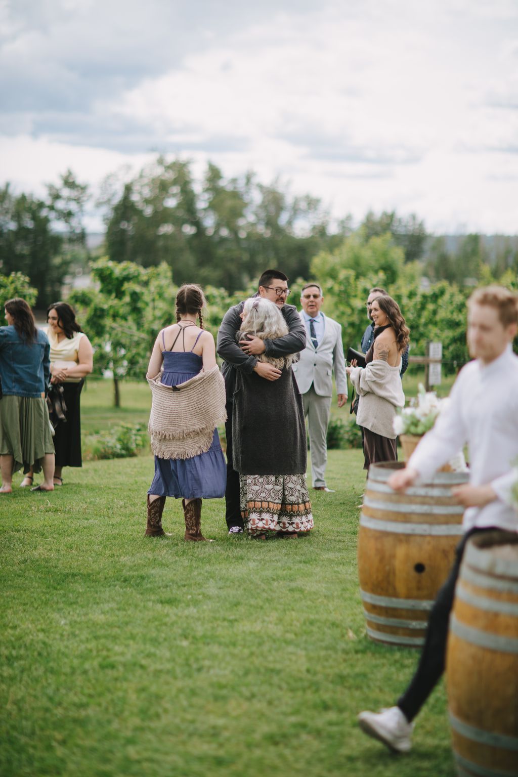 Groom embraces elderly woman during outdoor ceremony in vineyard with guests watching under cloudy sky
