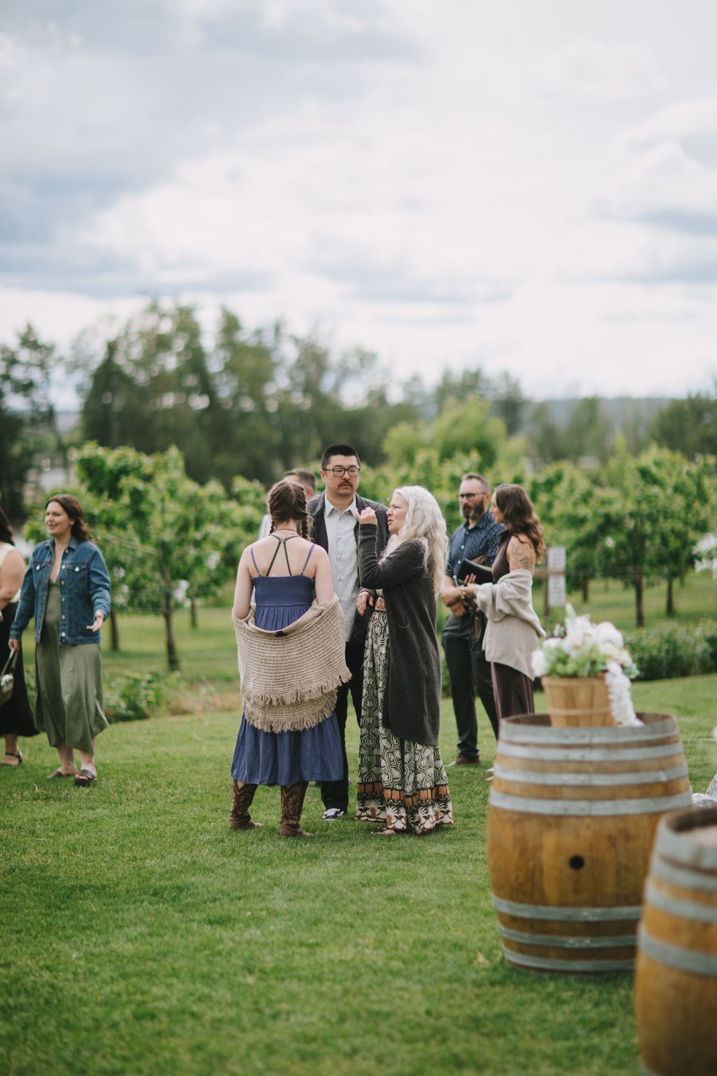 Bride in blue dress with cream shawl stands with groom in glasses during outdoor vineyard ceremony with guests and wooden barrels visible