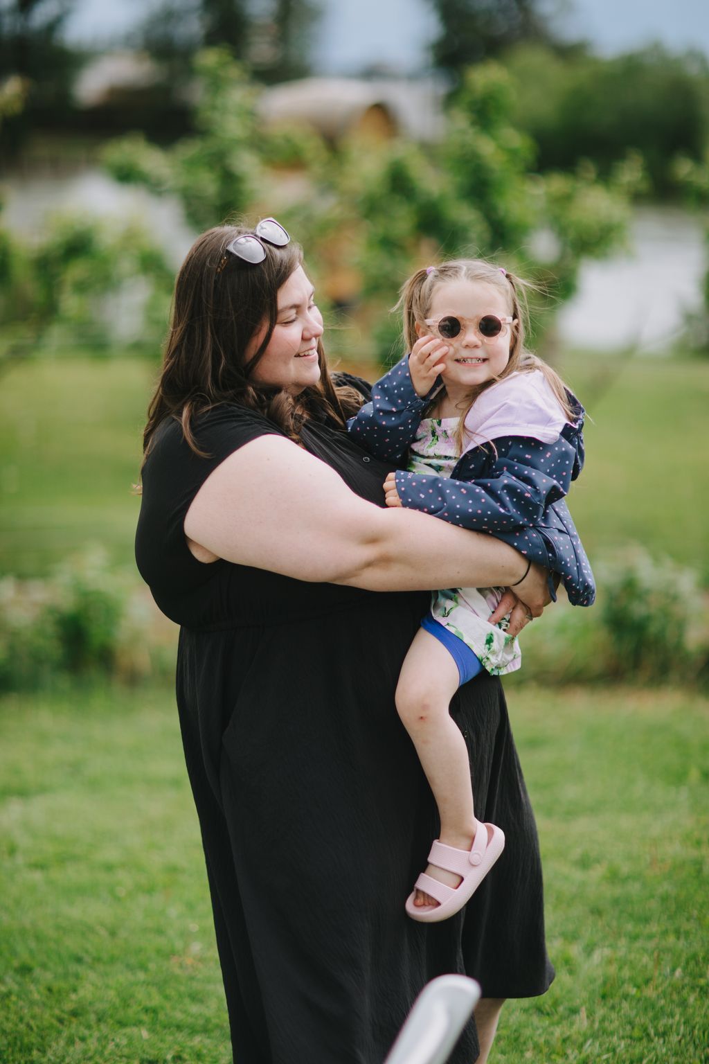 Woman holding young child in black dress outdoors, both wearing sunglasses, smiling in sunlit garden setting