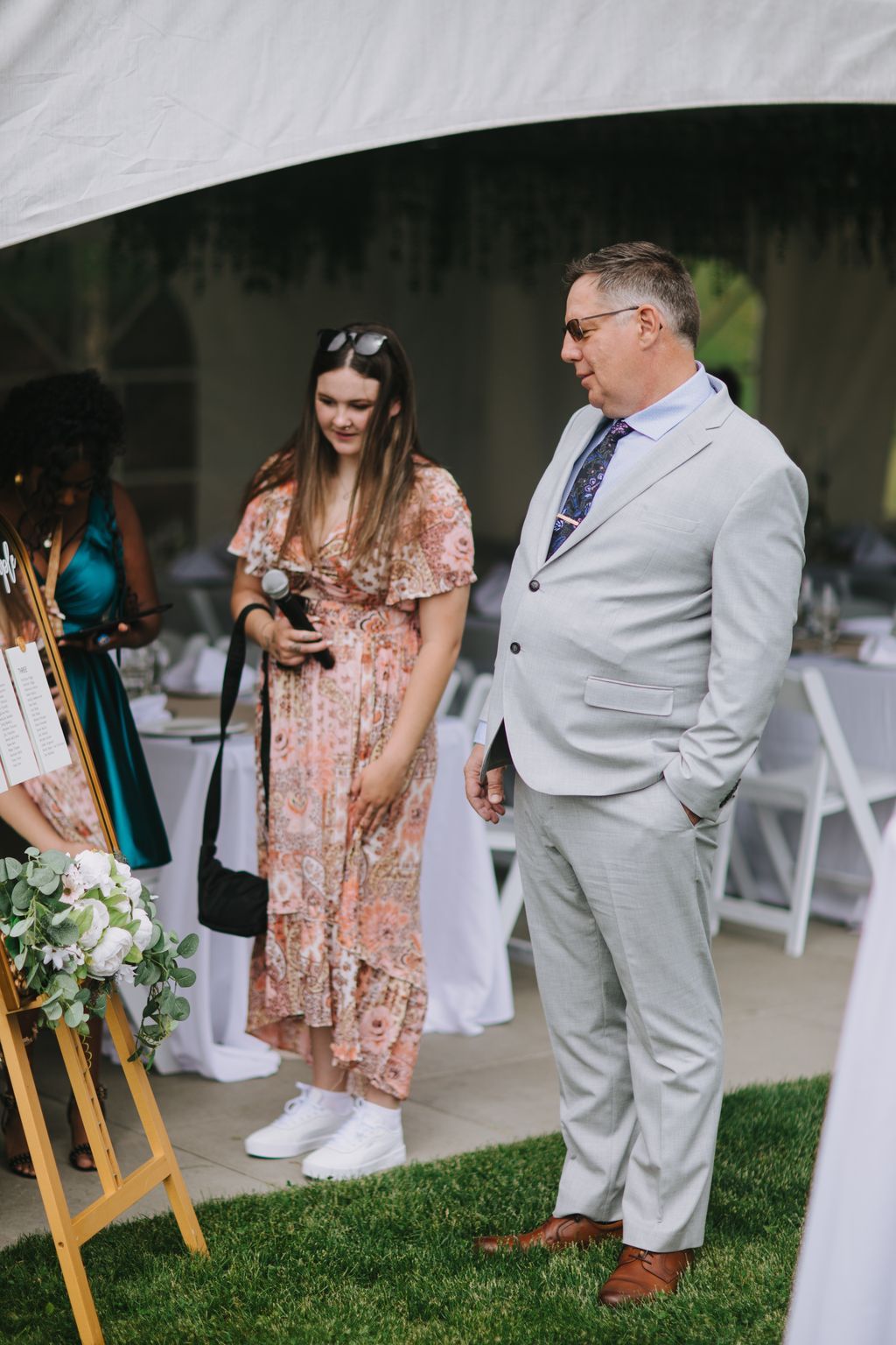 Bride in coral floral dress and groom in light gray suit stand together under white tent with artificial grass, exchanging smiles during outdoor wedding cere…